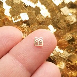 May include: A tiny gold-colored gift box charm with a bow on top. The charm is held between two fingers against a background of gold glitter. This charm is ideal for jewelry making or other craft projects.