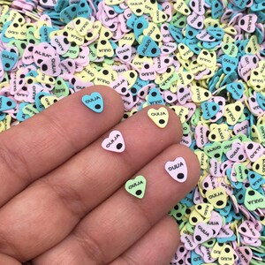 May include: A hand holding three small heart-shaped charms with the word "OUIJA" printed on them. The charms are green, pink, and blue. The background is a large pile of similar charms in various colors.