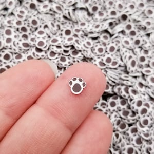 Puede incluir: Primer plano de una mano sosteniendo una pequeña huella de pata blanca con detalles negros. La huella de pata está rodeada de una gran cantidad de huellas de pata similares.