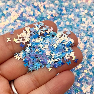 May include: A handful of iridescent blue butterfly-shaped glitter. The glitter is scattered on a white surface.