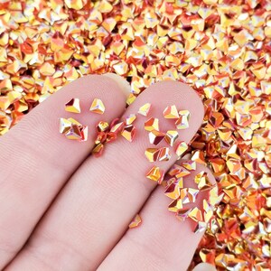 May include: A close-up of a hand holding a pile of iridescent orange and gold triangular glitter. The glitter is scattered on a surface.