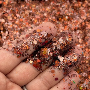 May include: A close-up of a hand holding a variety of glittery, iridescent, and metallic orange, brown, and copper colored glitter. The glitter is in a variety of shapes and sizes.