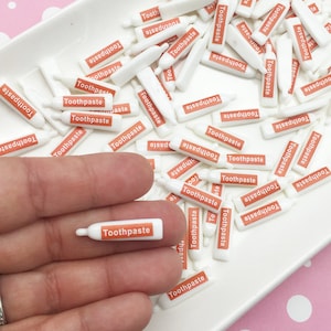 以下が含まれることがあります： オレンジ色の「Toothpaste」のラベルが付いた、ミニチュアの白い歯磨き粉チューブのコレクション。白い長方形の皿に並べられ、1本は手に持たれています。背景は白い水玉模様のピンク色の表面です。
