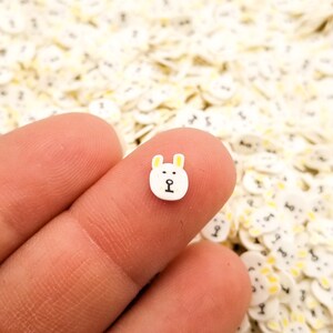 May include: A close-up of a white bear face with yellow ears, a black nose, and black eyes. The bear face is a small, flat charm, and there are many other similar charms in the background.
