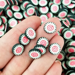 May include: A hand holding a variety of small, round, black and white cake decorations with a pink swirl design. The decorations are scattered on a surface.