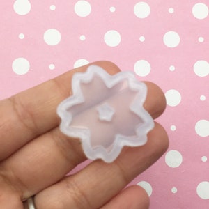 May include: A clear silicone mold shaped like a five-petal flower with a small circle in the center. The mold is held in a hand against a pink background with white polka dots.