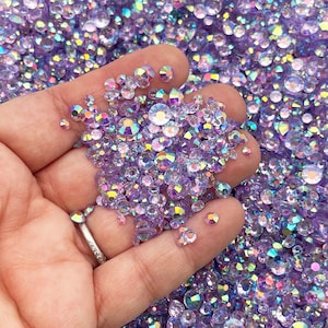 Puede incluir: Una mano sosteniendo una gran cantidad de piedras de cristal de color púrpura iridiscente y transparente. Las piedras de cristal son pequeñas y tienen un acabado facetado.