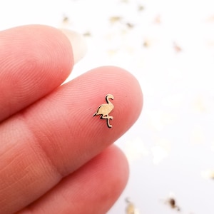 以下が含まれることがあります： 2 本の指で挟まれた、小さな金色メタルのフラミンゴの切り抜き。フラミンゴは右を向いている。