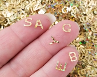 Puede incluir: Una mano sosteniendo pegatinas de letras doradas que deletrean "S A G E B". Las letras están sobre un fondo de muchas pegatinas de letras doradas.