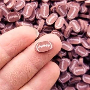 May include: A close-up of a hand holding a small brown and white football-shaped charm. The charm is surrounded by a large quantity of similar charms.