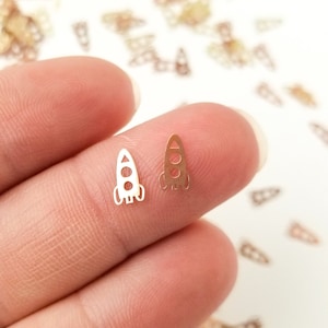 May include: Two small rocket ship shaped metal charms, one gold and one silver, held in a hand against a white background. The charms are surrounded by many smaller gold rocket ship charms.