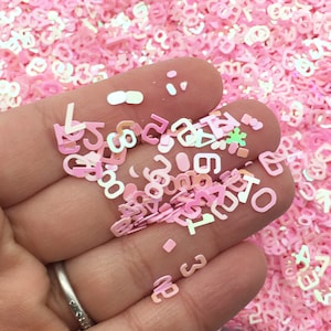 May include: A close-up of a hand holding pink iridescent letter and number shaped glitter. The glitter is scattered on a pink surface.