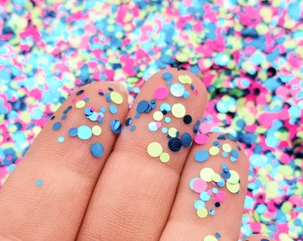 May include: A hand holding a variety of colorful glitter in shades of blue, green, and pink. The glitter is in the shape of small circles.