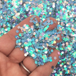 May include: A close-up of a hand holding a handful of iridescent blue glitter. The glitter is in various shapes and sizes, and it is very sparkly.