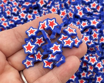 Op de afbeelding: Een hand die een stapel blauwe en rode stervormige polymer clay charms vasthoudt. De sterren hebben een witte omtrek en zijn verspreid over een oppervlak.