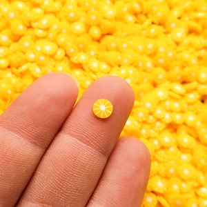 May include: A tiny, yellow and white lemon slice-shaped embellishment rests on a fingertip, set against a background of similar yellow shapes. The miniature detail suggests a craft or decorative item, possibly for nail art or scrapbooking.
