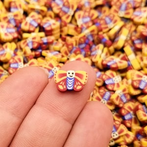 May include: A close-up of a hand holding a small, yellow, red, and blue butterfly charm. The charm has a smiling face and is surrounded by many other similar charms.