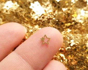 May include: A single gold star-shaped sequin is held between two fingers. The star is small and delicate, with a slightly textured surface. The background is a blurry image of a large quantity of gold star-shaped sequins.