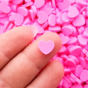 May include: A hand holding a single pink heart-shaped sprinkle against a background of many pink heart-shaped sprinkles.