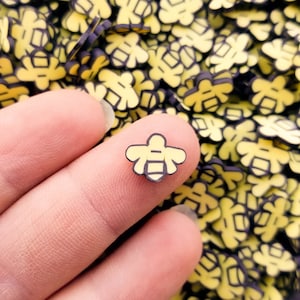 May include: A close-up of a finger holding a small yellow and black bee charm. The bee charm is surrounded by a large quantity of similar bee charms.