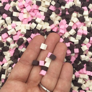May include: A hand holding a variety of small, colorful, square-shaped beads. The beads are pink, white, and brown. The beads are scattered on a surface.