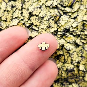 May include: A close-up of a yellow and black bee-shaped charm. The charm is being held by a hand against a background of many similar charms.