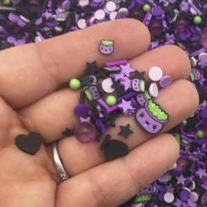 May include: A hand holding a variety of purple, black and green Halloween-themed sprinkles. The sprinkles are in the shape of stars, hearts, cauldrons and other fun designs.