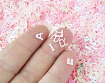 May include: A hand holding white plastic letters that spell out "A, I, R, E" against a background of pink iridescent letter confetti.
