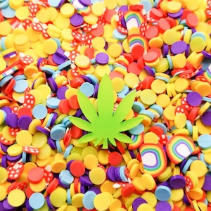 May include: A green marijuana leaf surrounded by colourful sprinkles, including yellow, red, blue, and purple. The sprinkles are in the shape of circles, rainbows, and mushrooms.
