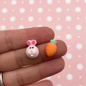 May include: Two small clay charms, one is a pink bunny with white eyes and a white nose, the other is an orange carrot with green leaves.