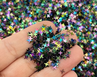 Pode incluir: Close-up de lantejoulas iridescentes em forma de estrela em tons de verde, roxo e azul. As lantejoulas estão espalhadas, criando um efeito brilhante. Estes enfeites decorativos são ideais para artesanato e projetos DIY.