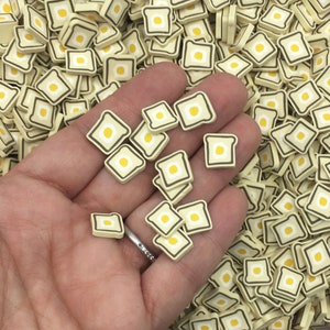 May include: A large pile of small, yellow and brown, square-shaped resin charms with a white egg design. The charms are in the shape of a piece of toast with a fried egg on top.