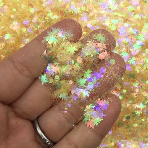 May include: Close-up of a hand holding a collection of glitter. The glitter features maple leaf shapes in shades of yellow, green, purple, and pink. The background is a light yellow color, and the glitter is scattered throughout.