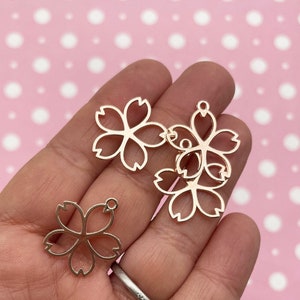May include: Four gold-colored metal charms shaped like cherry blossoms. The charms have a hole at the top for attaching to jewelry or other crafts.