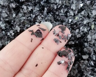May include: A hand holding a pile of black and silver iridescent glitter. The glitter is small and has a metallic sheen.