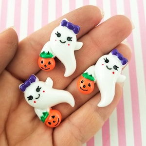 May include: Three white ghost charms with purple bows and orange pumpkin accents. The ghosts are holding small orange pumpkins with green stems. The charms are on a pink and white striped background.