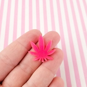 May include: A bright pink glitter resin cannabis leaf charm. The charm is being held in a hand.