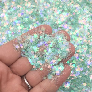 Op de afbeelding: Een close-up van iriserende groene glitter met verschillende vormen en maten. De glitter wordt in een hand gehouden, waardoor de textuur en glans zichtbaar worden.