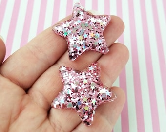 May include: Two pink glitter star-shaped embellishments. The stars are made of a clear resin material with pink glitter embedded in it. The glitter has a holographic finish, which gives it a rainbow effect.
