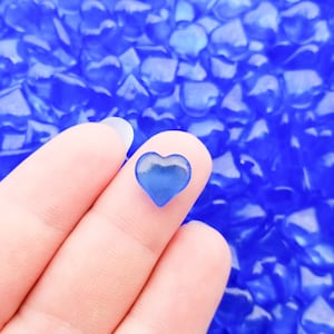May include: A single blue glass heart charm held between two fingers. The heart is small and translucent, with a slight shimmer. The background is a field of many smaller blue glass hearts.