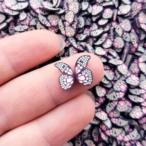 May include: A close-up of a hand holding a pink and black butterfly-shaped charm. The charm is surrounded by a large quantity of similar charms.
