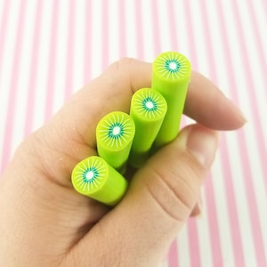 May include: Four green and white polymer clay canes with a kiwi fruit design. The canes are held in a hand.