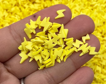 May include: A close-up of a hand holding a pile of yellow lightning bolt shaped sprinkles. The sprinkles are small and perfect for decorating cakes, cupcakes, and other desserts.