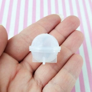May include: A clear, spherical mold for resin casting, held in a hand. The mold is composed of two halves that fit together, with a small square base. The background is a pink and white striped pattern.