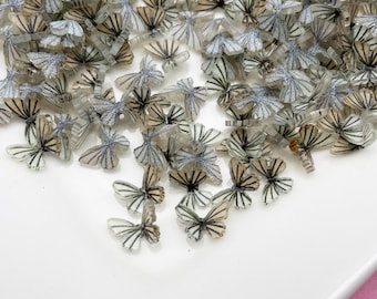 May include: A pile of clear plastic butterfly charms with black and silver glitter accents. The charms are scattered on a white surface.