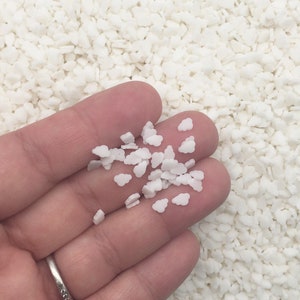 May include: A hand holding a small pile of white cloud-shaped sprinkles. The background is filled with similar white, irregular-shaped sprinkles. A silver ring is visible on the person's finger.