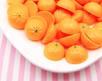 May include: A white plate holds numerous orange, miniature, citrus-shaped decorations. Some are whole, others halved, with a green dot at the top. The plate sits on a pink and white striped background. These decorations are likely for crafts or embellishments.