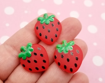 May include: Three red strawberry-shaped embellishments with green stems and black seeds. The strawberries are held in a hand against a pink background with white polka dots. These are likely craft supplies.