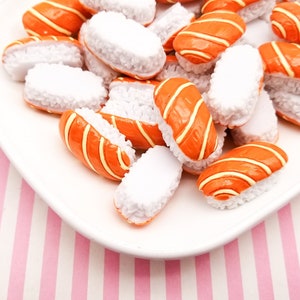 Könnte beinhalten: Ein weißer Teller gefüllt mit Miniatur-Sushi-Rollen. Die Rollen bestehen aus weißem Reis und haben eine dünne Schicht orangefarbenen Fisches auf der Oberseite. Der Teller befindet sich auf einer rosa-weißen gestreiften Oberfläche.