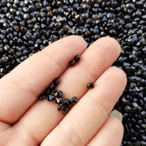 May include: A hand holding a small pile of black diamond-shaped beads. The beads are scattered on a surface covered in more of the same beads.